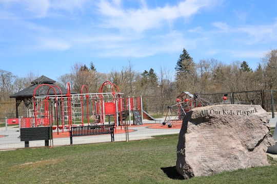 A playground with vibrant red and silver equipment, including climbing structures and slides, set within a fenced area. There is a large rock with an inscription marking the playpark. Surrounding the playground are trees and grass, and the sky is clear with a few clouds.