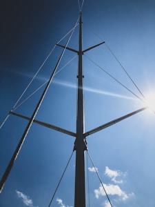A tall, vertical sailboat mast with a few spreaders extends into a clear blue sky. Thin lines, likely rigging, are attached to the mast and stretch out in different directions. The sun shines brightly from the right side, casting minimal shadows and enhancing the vibrancy of the blue sky with a few scattered white clouds.