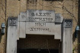 An entrance to an old brick building with an inscription reading 'Massie Township High School' in art deco style. Two lantern-style light fixtures are mounted on either side of the entrance. The building features decorative stone elements and appears to have some ivy or plants growing around it.