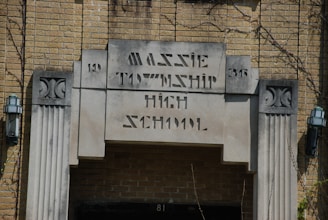 An entrance to an old brick building with an inscription reading 'Massie Township High School' in art deco style. Two lantern-style light fixtures are mounted on either side of the entrance. The building features decorative stone elements and appears to have some ivy or plants growing around it.