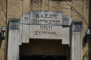 An entrance to an old brick building with an inscription reading 'Massie Township High School' in art deco style. Two lantern-style light fixtures are mounted on either side of the entrance. The building features decorative stone elements and appears to have some ivy or plants growing around it.