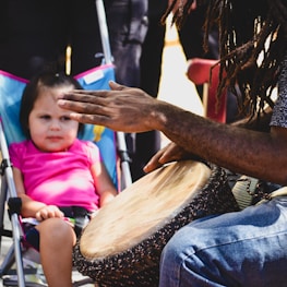 A young child wearing a pink shirt sits in a stroller, observing an individual playing a hand drum. The person playing the drum has long, braided hair and is wearing jeans. They are positioned close to the child, who appears intrigued by the drumming.