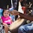 A young child wearing a pink shirt sits in a stroller, observing an individual playing a hand drum. The person playing the drum has long, braided hair and is wearing jeans. They are positioned close to the child, who appears intrigued by the drumming.
