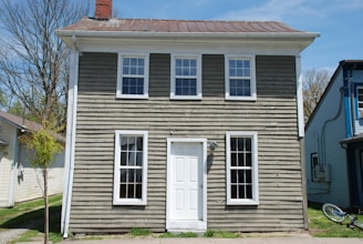 A small, two-story house with weathered wooden siding and a metal roof. The building has four windows on the front, two on each floor, and a white door in the center. The house is flanked by neighboring buildings, and a small tree is visible on the left side. A bicycle is leaning against the side of the house.