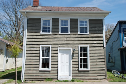 A small, two-story house with weathered wooden siding and a metal roof. The building has four windows on the front, two on each floor, and a white door in the center. The house is flanked by neighboring buildings, and a small tree is visible on the left side. A bicycle is leaning against the side of the house.