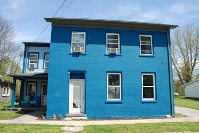 A two-story house painted bright blue with multiple windows is captured. The front door is white and there's a small porch with two blue chairs. The surroundings include a lawn and some trees, with another house visible in the background.