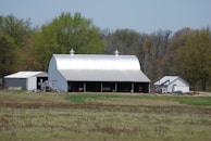 A large, metallic barn with a curved roof is situated within a rural setting, surrounded by lush green trees. Adjacent to it are smaller utility buildings and scattered farming equipment. The foreground features a field with patches of grass and earthy areas, indicating agricultural land.