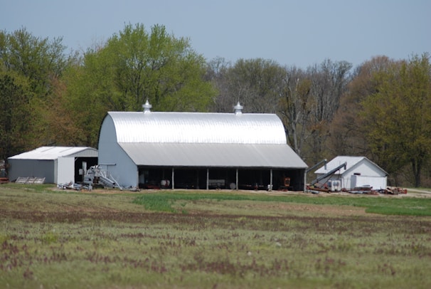A large, metallic barn with a curved roof is situated within a rural setting, surrounded by lush green trees. Adjacent to it are smaller utility buildings and scattered farming equipment. The foreground features a field with patches of grass and earthy areas, indicating agricultural land.