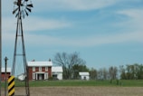 A rural scene featuring a red brick farmhouse with a white roof, surrounded by fields and trees. A traditional windmill tower stands in the foreground, to the left, and a barn with a similar white roof is seen in the background. The sky is mostly clear with a few wispy clouds.