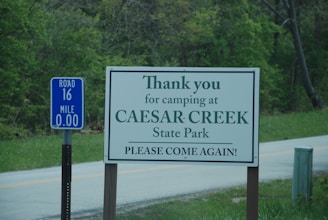 A sign positioned by the roadside expresses gratitude to visitors for camping at Caesar Creek State Park, encouraging them to return. Another blue sign beside it marks Road 16 and Mile 0.00. The surrounding area features lush green trees and a small paved road.