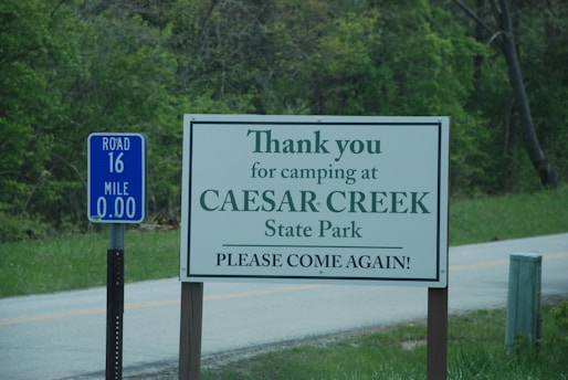 A sign positioned by the roadside expresses gratitude to visitors for camping at Caesar Creek State Park, encouraging them to return. Another blue sign beside it marks Road 16 and Mile 0.00. The surrounding area features lush green trees and a small paved road.