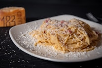A plate of spaghetti carbonara topped with a generous amount of grated cheese sits on a square white plate. In the background, a wedge of cheese with marked numbers is visible, alongside a fork on the right side. The setting is dimly lit, creating a cozy and appetizing atmosphere.