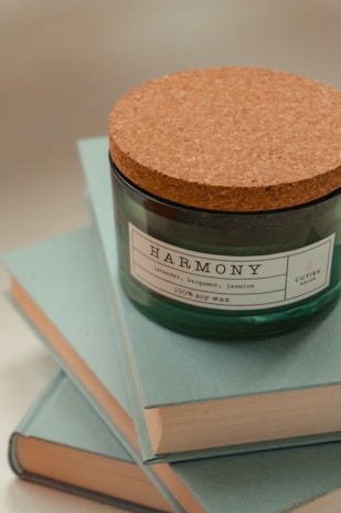 A green candle labeled 'Harmony' with scents of lavender, bergamot, and jasmine. The candle has a cork lid and rests on a stack of light blue books.