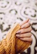 A hand is shown with elegantly manicured nails, featuring a glittery accent on one finger. The nails are embellished with small decorative gems. The hand is partially covered by a mustard-colored knitted sweater. The background consists of a soft, blurred abstract pattern.