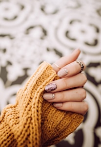 A hand is shown with elegantly manicured nails, featuring a glittery accent on one finger. The nails are embellished with small decorative gems. The hand is partially covered by a mustard-colored knitted sweater. The background consists of a soft, blurred abstract pattern.