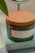 A glass candle jar with a cork lid is placed on a stack of light blue books. The jar label reads 'HARMONY', featuring scents such as lavender, bergamot, and jasmine, and indicates it contains 100% soy wax. A green leaf can be seen on the left side of the image.