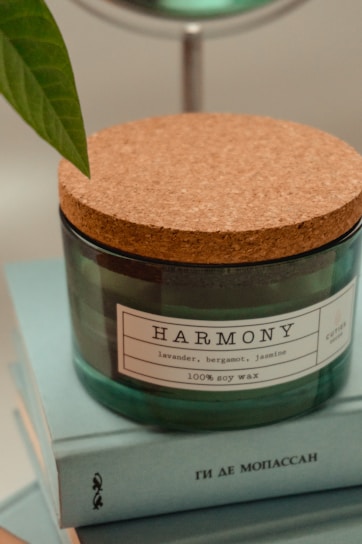 A glass candle jar with a cork lid is placed on a stack of light blue books. The jar label reads 'HARMONY', featuring scents such as lavender, bergamot, and jasmine, and indicates it contains 100% soy wax. A green leaf can be seen on the left side of the image.