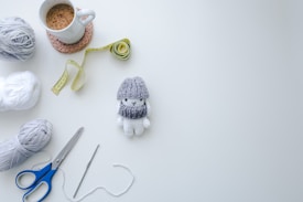 A flat lay arrangement with a cozy, hand-crafted feel includes a mug of coffee on a crocheted coaster, a yellow measuring tape, a handmade knitted doll wearing a gray hat and scarf, balls of gray and white yarn, a pair of scissors with blue handles, and a crochet hook with attached yarn against a white background.