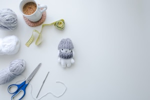 A flat lay arrangement with a cozy, hand-crafted feel includes a mug of coffee on a crocheted coaster, a yellow measuring tape, a handmade knitted doll wearing a gray hat and scarf, balls of gray and white yarn, a pair of scissors with blue handles, and a crochet hook with attached yarn against a white background.