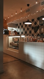 A cozy indoor setting combining modern and rustic styles, featuring a checkered wall and a tiled counter. There are various bottles and a clock mounted on the wall. A display case shows a selection of pastries and bread, and soft lighting enhances the warm atmosphere.