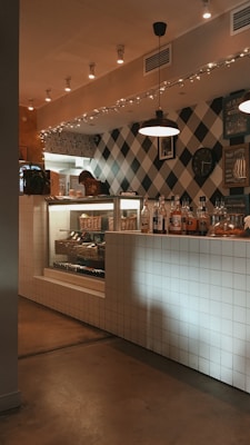 A cozy indoor setting combining modern and rustic styles, featuring a checkered wall and a tiled counter. There are various bottles and a clock mounted on the wall. A display case shows a selection of pastries and bread, and soft lighting enhances the warm atmosphere.