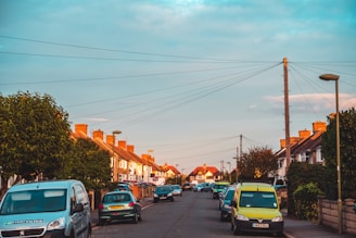 A suburban street lined with cars parked along both sides. The houses are traditional, with red brick chimneys and sloping roofs. Trees and bushes are visible, and streetlights dot the landscape. Overhead, numerous power lines crisscross between utility poles. The sky is clear with soft clouds, suggesting a calm and pleasant day.