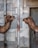 Two camels face each other, appearing to be in a conversation or intimate encounter. They are situated against a backdrop of a textured, industrial wall made of square stone tiles. A rusty metal pole divides the two camels in the center of the image.