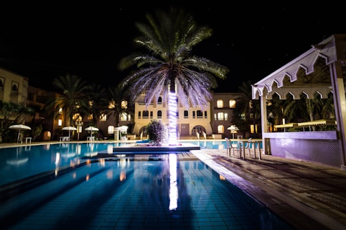 Luxurious pool area at dusk, illuminated with subtle motion lights reflecting off the water and surrounded by palm trees.