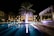A luxurious, well-lit swimming pool area at night surrounded by a few buildings. Palm trees are prominently illuminated, casting reflections on the water. Chairs and tables are placed near the poolside under a canopy.