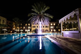 A luxurious, well-lit swimming pool area at night surrounded by a few buildings. Palm trees are prominently illuminated, casting reflections on the water. Chairs and tables are placed near the poolside under a canopy.