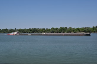 A cargo boat navigating a calm river surrounded by lush greenery under a clear sky.