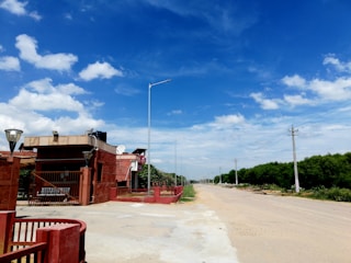 A expansive road stretches into the distance alongside a series of utility poles and lush green trees under a vibrant blue sky with scattered white clouds. A brick-red building featuring multiple satellite dishes and lamps stands in the foreground.