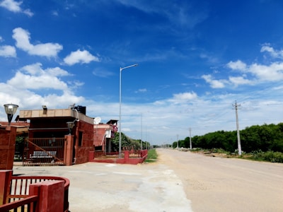 A expansive road stretches into the distance alongside a series of utility poles and lush green trees under a vibrant blue sky with scattered white clouds. A brick-red building featuring multiple satellite dishes and lamps stands in the foreground.