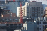 Urban cityscape with multiple buildings of varying heights and architectural styles. A prominent red construction crane is seen in the midst of mid-rise buildings, indicating ongoing construction in the area. The buildings in the background have modern glass façades, while those in the foreground appear more utilitarian with fewer windows. The scaffolding and construction materials on the lowermost buildings suggest further development.