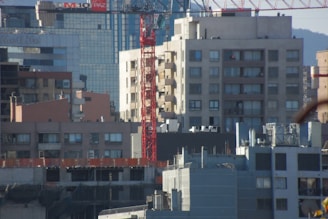 Urban cityscape with multiple buildings of varying heights and architectural styles. A prominent red construction crane is seen in the midst of mid-rise buildings, indicating ongoing construction in the area. The buildings in the background have modern glass façades, while those in the foreground appear more utilitarian with fewer windows. The scaffolding and construction materials on the lowermost buildings suggest further development.