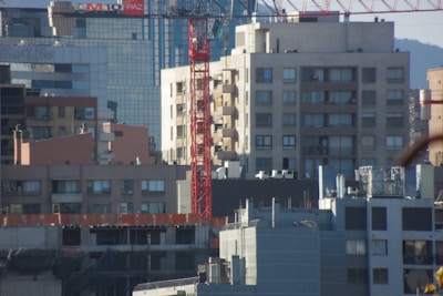 Urban cityscape with multiple buildings of varying heights and architectural styles. A prominent red construction crane is seen in the midst of mid-rise buildings, indicating ongoing construction in the area. The buildings in the background have modern glass façades, while those in the foreground appear more utilitarian with fewer windows. The scaffolding and construction materials on the lowermost buildings suggest further development.