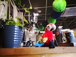 A colorful parrot plush toy sits on a wooden surface next to a blue plant pot and a glass coffee siphon. Above the table, paper decorations in shades of green and blue hang from the ceiling. There are also paper cups with floral designs on the surface.