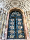 A grand, ornate door with intricate gold and blue metalwork set within a richly decorated archway. The arch features detailed carvings and stone columns on both sides, showcasing a blend of textures and historical design elements.