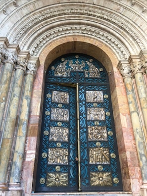 A grand, ornate door with intricate gold and blue metalwork set within a richly decorated archway. The arch features detailed carvings and stone columns on both sides, showcasing a blend of textures and historical design elements.