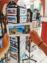 A person is holding a postcard depicting a scenic beach view with cliffs and blue water. The setting appears to be an outdoor postcard stand with multiple racks filled with postcards featuring various travel destinations.