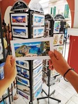 A person is holding a postcard depicting a scenic beach view with cliffs and blue water. The setting appears to be an outdoor postcard stand with multiple racks filled with postcards featuring various travel destinations.