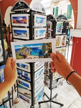 A person is holding a postcard depicting a scenic beach view with cliffs and blue water. The setting appears to be an outdoor postcard stand with multiple racks filled with postcards featuring various travel destinations.