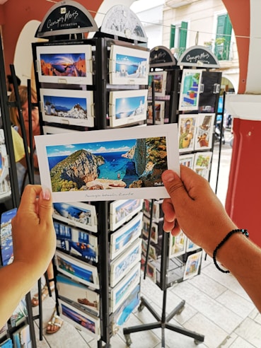 A person is holding a postcard depicting a scenic beach view with cliffs and blue water. The setting appears to be an outdoor postcard stand with multiple racks filled with postcards featuring various travel destinations.