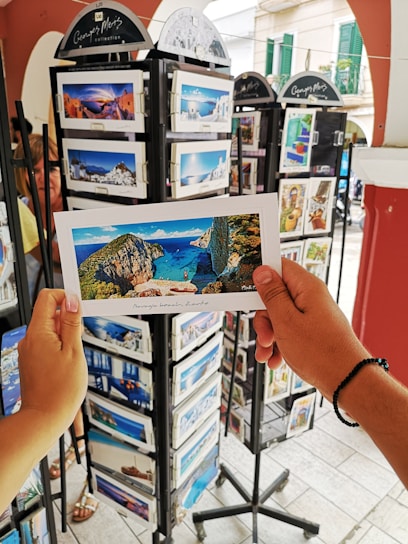 A person is holding a postcard depicting a scenic beach view with cliffs and blue water. The setting appears to be an outdoor postcard stand with multiple racks filled with postcards featuring various travel destinations.