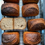 Several loaves of freshly baked bread are arranged on a cooling rack. The bread has a rich, golden-brown crust with visible seeds on the surface. One loaf is sliced open to reveal a soft, airy interior with an even crumb structure. The arrangement gives a sense of artisanal baking.