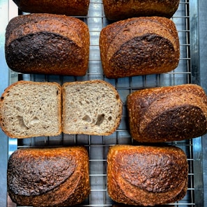 Several loaves of freshly baked bread are arranged on a cooling rack. The bread has a rich, golden-brown crust with visible seeds on the surface. One loaf is sliced open to reveal a soft, airy interior with an even crumb structure. The arrangement gives a sense of artisanal baking.