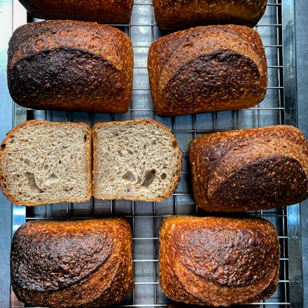 Several loaves of freshly baked bread are arranged on a cooling rack. The bread has a rich, golden-brown crust with visible seeds on the surface. One loaf is sliced open to reveal a soft, airy interior with an even crumb structure. The arrangement gives a sense of artisanal baking.