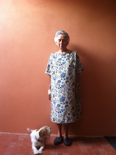 An elderly woman is standing against a plain, warm-colored wall. She is wearing a loose-fitting, floral-patterned dress in shades of blue and white. Next to her, a small white and brown dog is looking up at her. The woman has short gray hair and is wearing dark slippers.