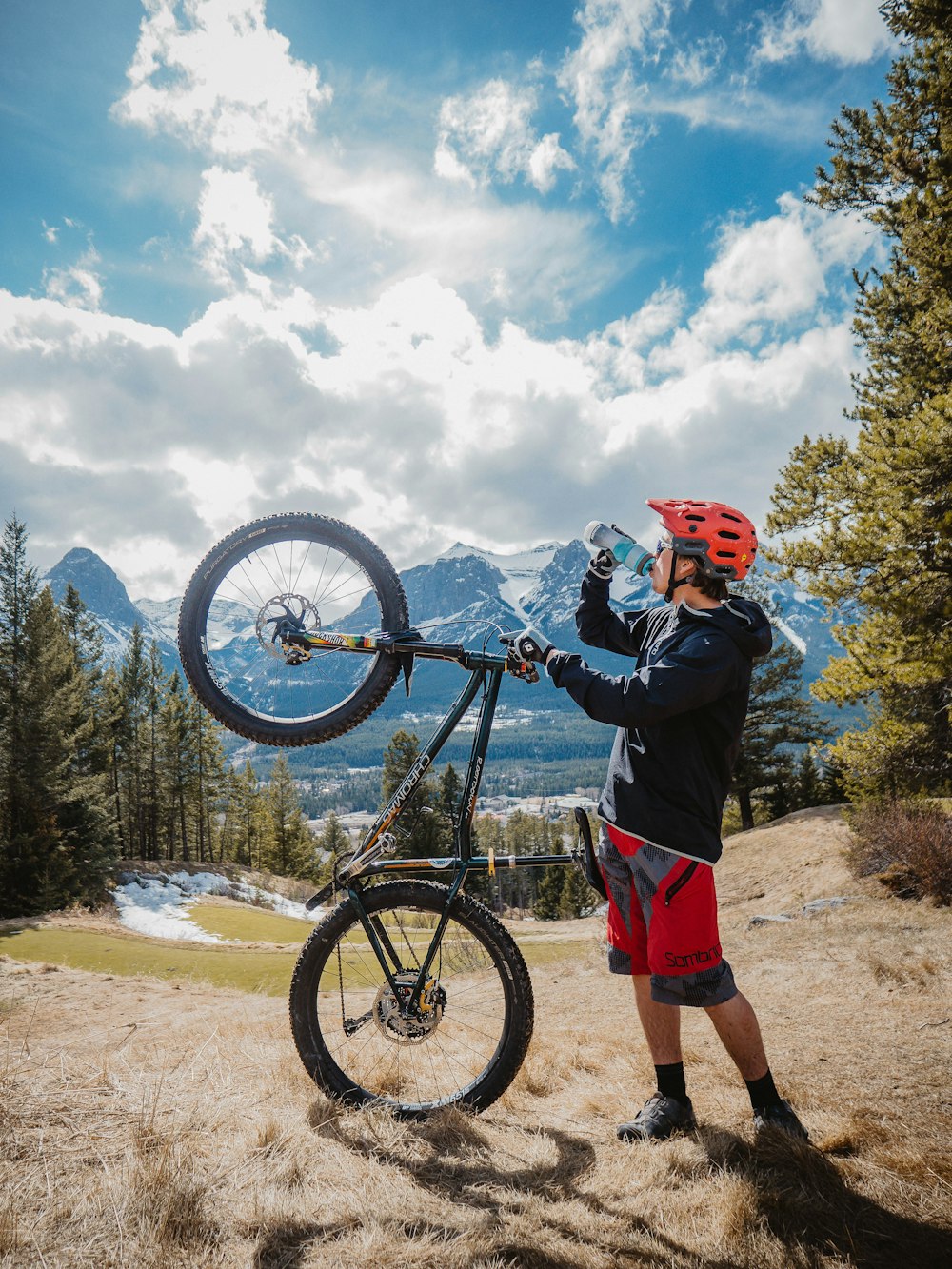 homme en veste bleue et short rouge debout à côté d’un vélo de montagne noir pendant la journée