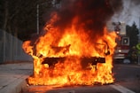 A car engulfed in intense flames on the side of a road with thick black smoke billowing into the air. Firefighters and a fire truck are visible in the background, indicating an emergency response situation.